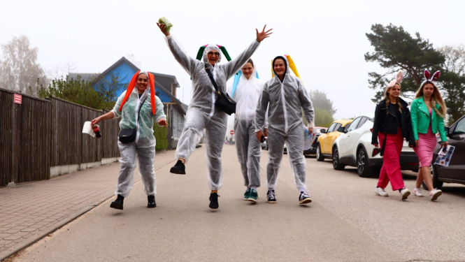 Cilvēku grupa jautrā noskaņojumā iet pa ielu, vairāki valkā caurspīdīgus lietusmēteļus un zaķa ausis.