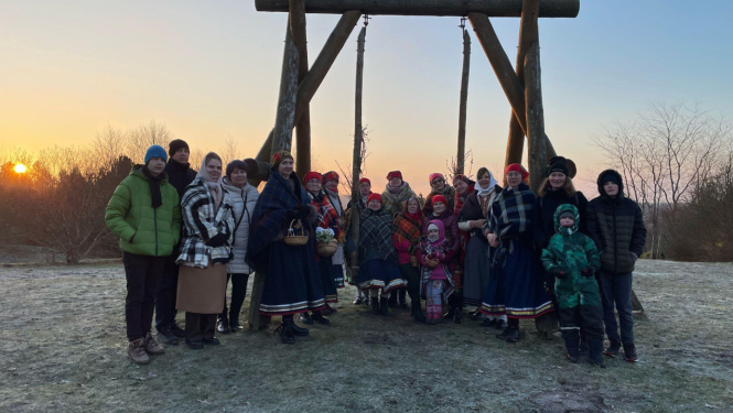 Cilvēku grupa ziemīgā ainavā pie koka šūpolēm saulrietā; daļa tērpusies tradicionālos tautastērpos, citi – siltā ikdienas apģērbā.