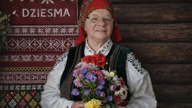 Attēlā Zenta Bērtiņa tautas tērpā ar vasaras ziedu pušķi.
