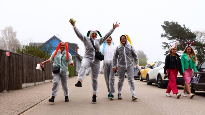 Cilvēku grupa jautrā noskaņojumā iet pa ielu, vairāki valkā caurspīdīgus lietusmēteļus un zaķa ausis.