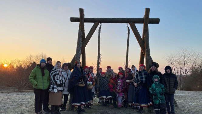 Cilvēku grupa ziemīgā ainavā pie koka šūpolēm saulrietā; daļa tērpusies tradicionālos tautastērpos, citi – siltā ikdienas apģērbā.