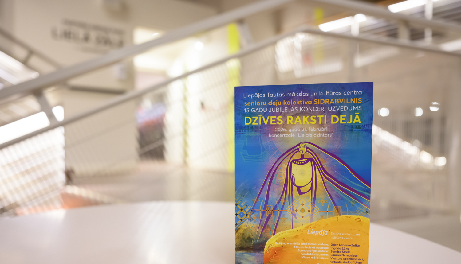 Uz galda novietots krāsains plakāts modernā, gaišā interjerā ar izplūdušu fonu