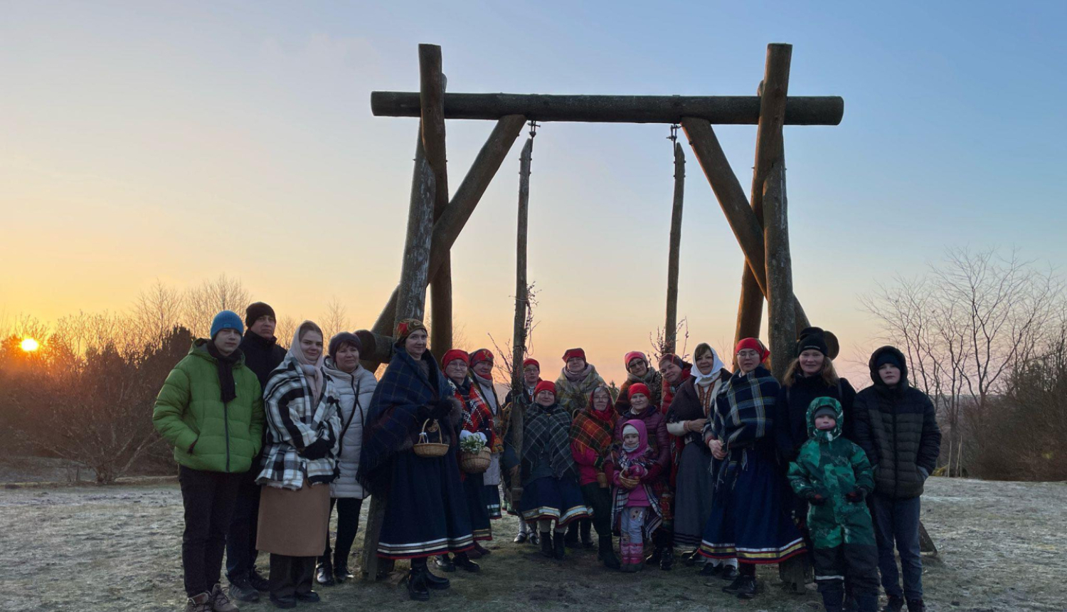 Cilvēku grupa ziemīgā ainavā pie koka šūpolēm saulrietā; daļa tērpusies tradicionālos tautastērpos, citi – siltā ikdienas apģērbā.