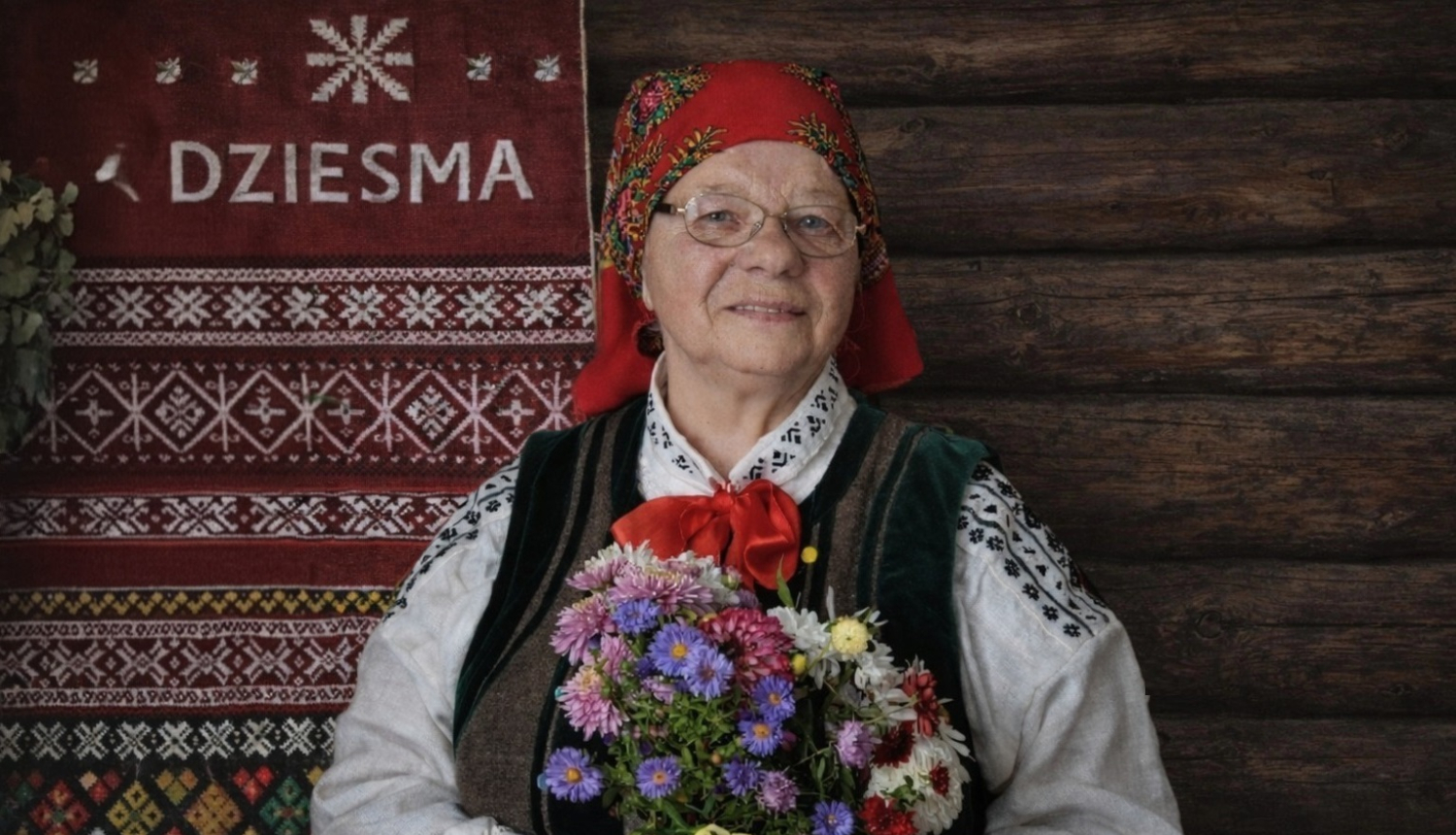 Attēlā Zenta Bērtiņa tautas tērpā ar vasaras ziedu pušķi.